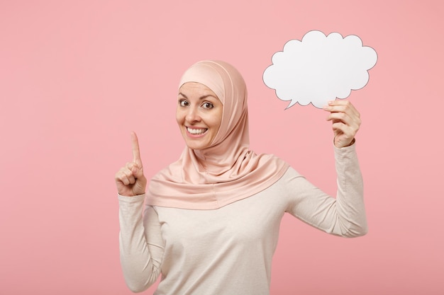 Arabska Muzułmanka w jasnych ubraniach hidżab pozowanie na białym tle na różowym tle. Koncepcja życia religijnego ludzi. Makieta miejsca na kopię. Trzymając pusty pusty Say cloud, wskazując palcem w górę z nowym pomysłem.