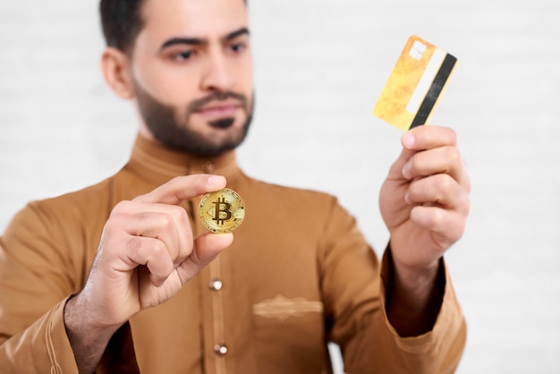 Arabski Biznesmen Trzyma Złoty Bitcoin W Jednej Ręce I Złotą Kartę Kredytową W Drugiej. Nosi Beżową Koszulę Z Wzorem. Zbliżenie Zostało Wykonane Na Białym Tle Studio.