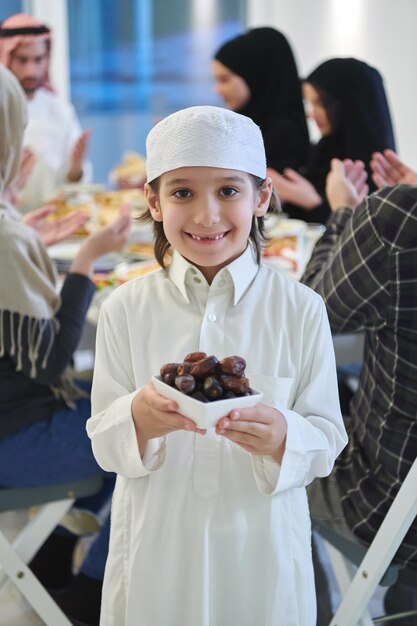 Arabski dzieciak w tradycyjnych strojach podczas iftar. Portret szczęśliwego młodego muzułmańskiego chłopca trzymającego miskę daktyli, podczas gdy reszta rodziny je