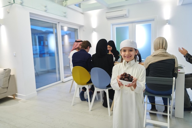 Arabski dzieciak w tradycyjnych strojach podczas iftar. Portret szczęśliwego młodego muzułmańskiego chłopca trzymającego miskę daktyli, podczas gdy reszta rodziny je