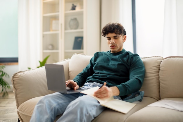 Arabski student pracujący z laptopem i robiący notatki w domu