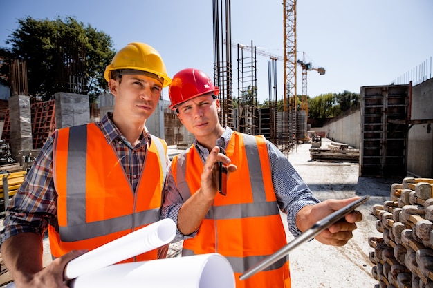 Architekt i inżynier budowlany ubrany w pomarańczowe kamizelki robocze i hełmy omawiają projekt budowlany na tablecie na placu budowy na świeżym powietrzu z materiałem budowlanym. .