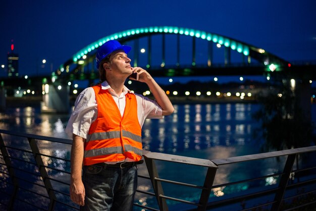 Architekt rozmawiający na telefonie komórkowym z oświetlonym mostem i światłami miasta w tle.