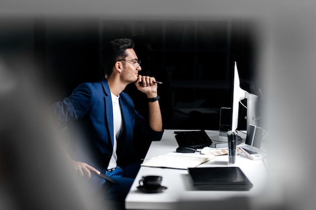 Architekt w okularach ubrany w niebieską marynarkę siedzi przy biurku przed komputerem i myśli.