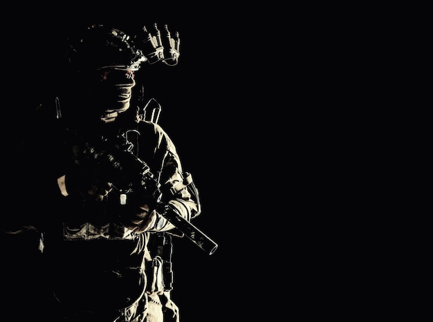 Armia operacji specjalnych sił żołnierz w masce i mundurze bojowym wyposażony w hełm wyposażony w noktowizor uzbrojony pistolet maszynowy z tłumikiem patrząc na bok niski klucz portret studyjny na czarnym copyspace