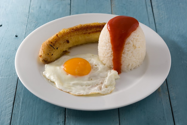 Arroz A La Cubana Typowy Kubański Ryż Ze Smażonym Bananem I Jajkiem Sadzonym Na Talerzu Na Drewnianym Stole.