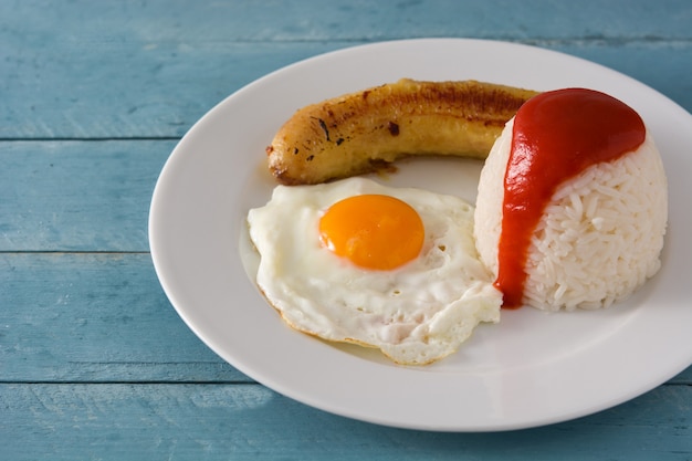 Arroz A La Cubana Typowy Kubański Ryż Ze Smażonym Bananem I Jajkiem Sadzonym Na Talerzu Na Drewnianym Stole.