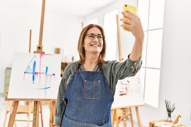 Artystka w średnim wieku kobieta uśmiecha się szczęśliwa robiąc selfie smartfonem w studio artystycznym.