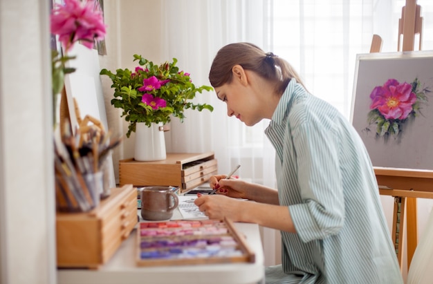 Artystka z boku rysuje bukiet róż i różowych piwonii przy stole w miejscu pracy, siedząc z pudełkami z kredkami suchych pasteli i materiałami do kreatywności. Koncepcja kreatywności i hobby