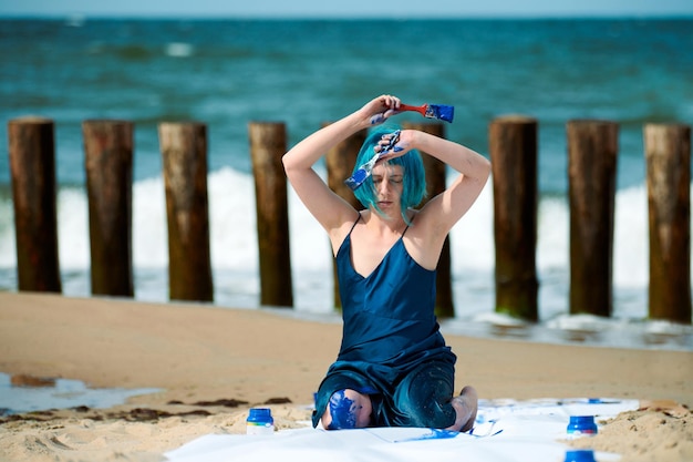 Artystyczna niebieskowłosa kobieta performer w granatowej sukience posmarowanej niebieskim gwaszem, siedząc na plaży. Koncepcja malowania ciała i sztuki ciała, ekspresyjna sztuka performance na świeżym powietrzu