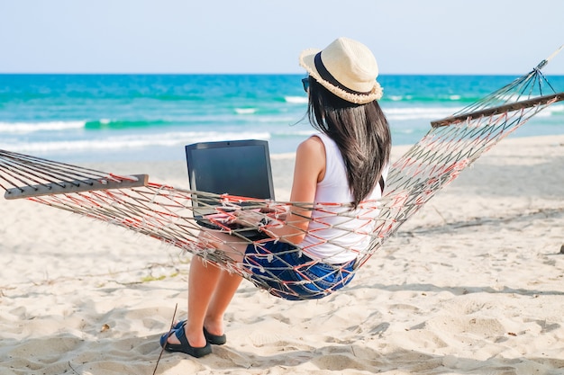 Zdjęcie asia kobiety obsiadanie na hamaku blisko plaży i używać laptop dla jej pracy i sprawdzać biznes podczas jej wakacji.