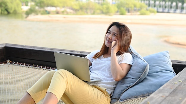 Asian businesswoman pracy online na plaży. Freelancer wykorzystujący technologię do pracy wszędzie. Kobieta rozmawia z klientem z telefonem komórkowym.