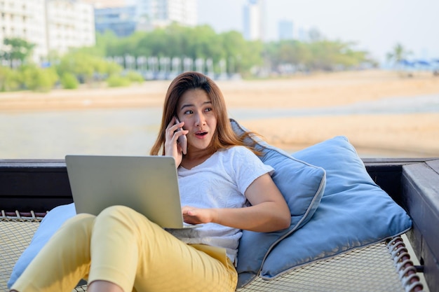 Asian businesswoman pracy online na plaży. Freelancer wykorzystujący technologię do pracy wszędzie. Kobieta rozmawia z klientem z telefonem komórkowym.