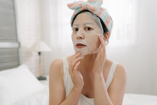 asian japonka w pałąk robi zabieg anty skazy skóry. Poranna pielęgnacja skóry. młoda kobieta ładny twarz aparat i zakładanie maski na twarz siedząc w białym wygodnym łóżku w domu.