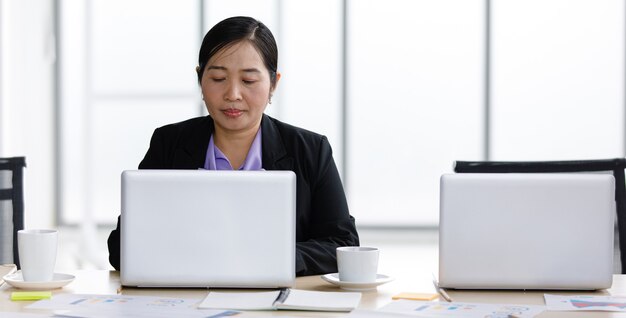 Asian średnim wieku szczęśliwa kobieta udana businesswoman sekretarka w formalnym garniturze siedzi przy biurku, wpisując informacje na laptopie komputer przenośny sam w cichym pomieszczeniu biurowym firmy.