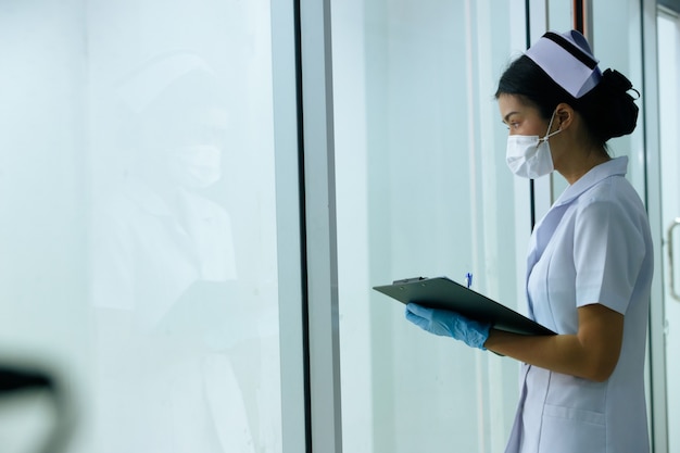Asian Woman Nurse sprawdzanie wykresu pacjenta jako rutynowe, pielęgniarka wygląda przez okna na zewnątrz oddziału opieki lub izby przyjęć w szpitalu. Koncepcja gościnności opieki zdrowotnej