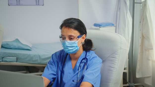 Asystent oddziału posiadający maskę ochronną i okulary, piszący raport na laptopie. Lekarz specjalista medycyny udzielający świadczeń zdrowotnych leczenie konsultacyjne w gabinecie szpitalnym w okresie Covid-19