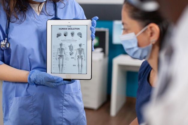 Asystentka Pediatry W Rękawiczkach Ochronnych Przed Covid19 Trzymająca Komputer Typu Tablet Z Radiografią Kości Na Ekranie. Afroamerykański Lekarz Wyjaśniający Wiedzę Medyczną W Gabinecie Szpitalnym