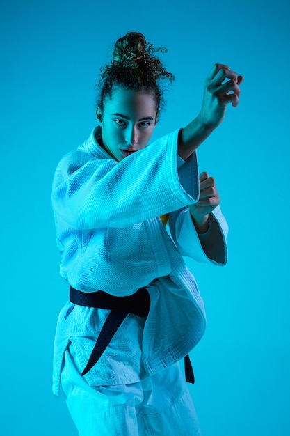 Atak. Profesjonalna judoistka w białym kimono judo praktykowania i szkolenia na białym tle na niebieskim tle studio neoned. Łaska ruchu i działania. Zdrowy styl życia, koncepcja sportu