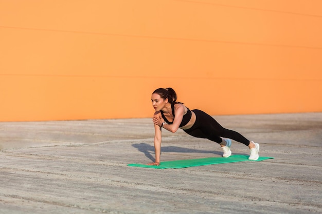 Atletyczna sprawna ładna dziewczyna w sportowej czarne spodnie i top ćwiczenie jogi stojący w pozie wyższej deski Phalakasana jedną ręką trening siły mięśni Opieka zdrowotna aktywność sportowa na świeżym powietrzu