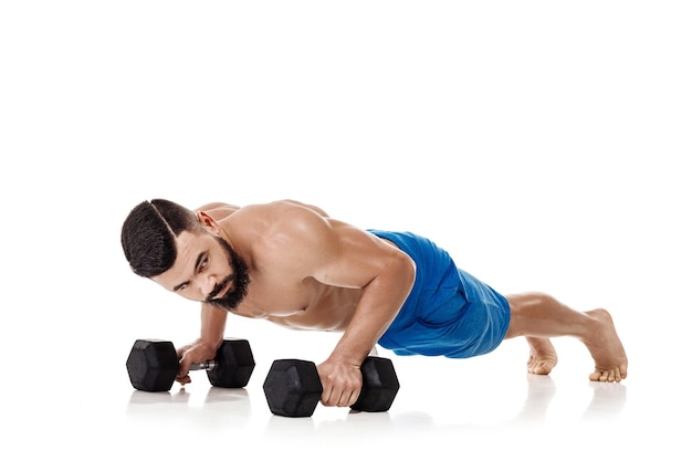 Atletyczny muskularny mężczyzna robi ćwiczenia push up z hantlami. Silny kulturysta z nagim torsem na białym tle