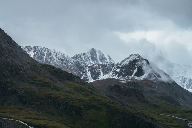 Atmosferyczny krajobraz alpejski z wielką górą szczytową ze śniegiem w niskich chmurach. Dramatyczna górska sceneria z ostrym szczytem w pochmurną pogodę. Niesamowity widok na śnieżny spiczasty szczyt w zachmurzonym niebie.