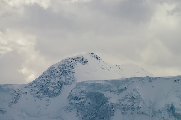 Atmosferyczny krajobraz góralski z wysokimi śnieżnymi górskimi ścianami pod zachmurzonym niebem. Dramatyczna sceneria z pokrytym śniegiem szczytem góry w pochmurną pogodę. Światło słoneczne przez chmury nad szczytem śniegu.