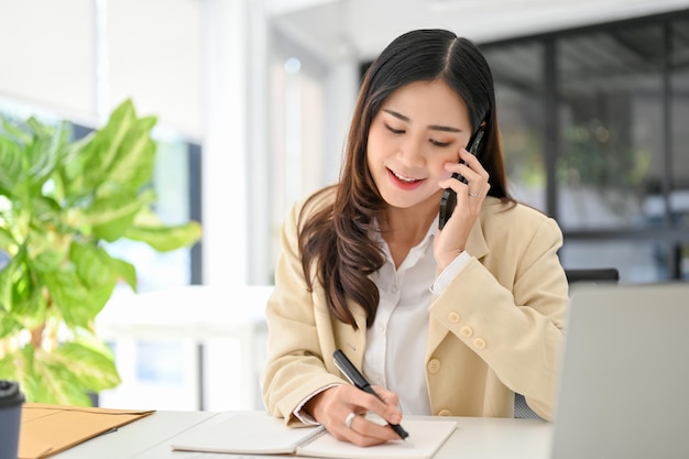 Atrakcyjna azjatycka bizneswoman robi notatki na swoim notatniku podczas rozmowy przez telefon
