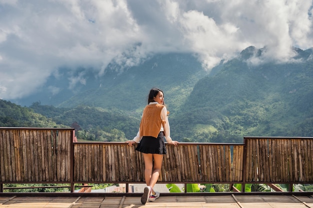 Atrakcyjna azjatycka kobieta stojąca i ciesząca się widokiem na góry na drewnianym balkonie w słoneczny dzień w Chiang Dao, Chiang Mai