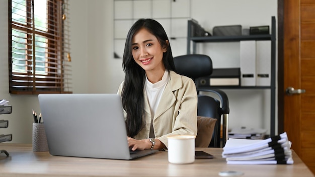 Atrakcyjna azjatycka kobieta w biurze siedzi przy biurku, uśmiechając się i patrząc na kamerę