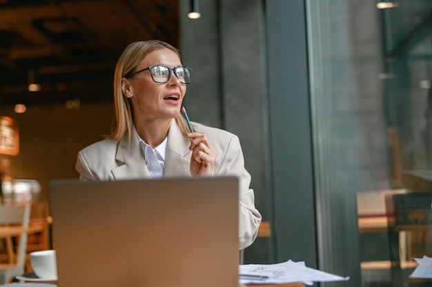 Atrakcyjna bizneswoman freelancer pracuje na laptopie siedząc w kawiarni przy oknie