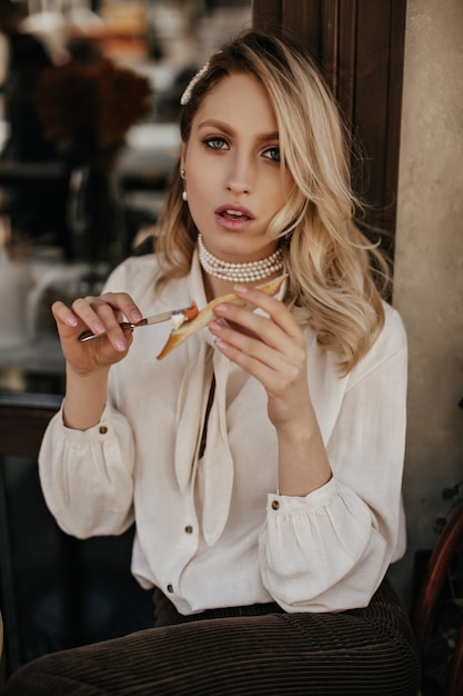 Atrakcyjna blondynka kręcone kobieta rozprowadza pastę na bruschetcie. Ładna pani w białej bluzce i aksamitnych spodniach siedzi w ulicznej kawiarni