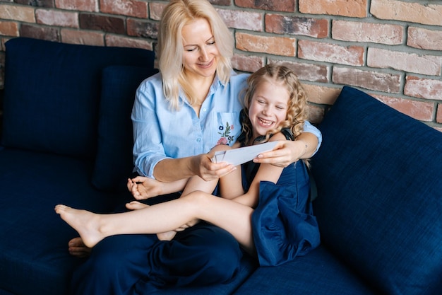Atrakcyjna blondynka w codziennych ubraniach i jej córeczka przeglądają zdjęcia i uśmiechają się siedząc na kanapie w domu w przytulnym salonie. Pojęcie rodzinnych wakacji i wspólnoty.