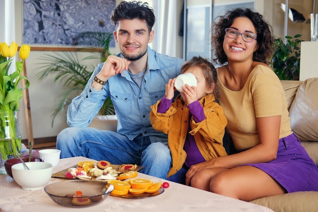 Atrakcyjna brunetka kobieta w okularach, brodaty stylowy mężczyzna i ich urocza córeczka siedzą na kanapie i jedzą obiad w salonie.