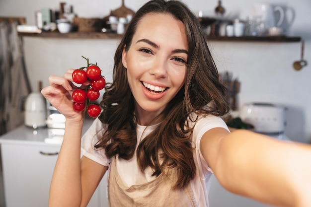 Atrakcyjna brunetka w fartuchu robi selfie siedząc w kuchni w domu, trzymając kiść pomidorów