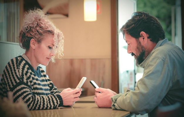 Atrakcyjna dojrzała para rasy kaukaskiej siedząca przy stole w restauracji za pomocą profesjonalnego modela wpływowego smartfona śledzącego media społecznościowe