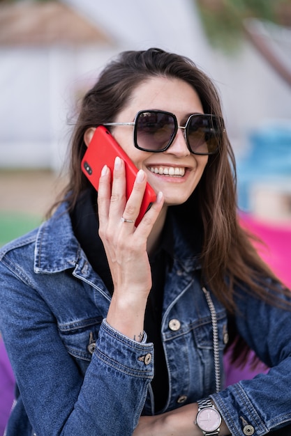 Atrakcyjna Dziewczyna Odpoczywa Na Naturze W Weekend. Dziewczyna W Dżinsowej Kurtce Siedzi W Parku. Kaukaska Brunetki Młoda Kobieta Opowiada Na Smartphone Na Zewnątrz Samotnie. Technologie I Gadżety.