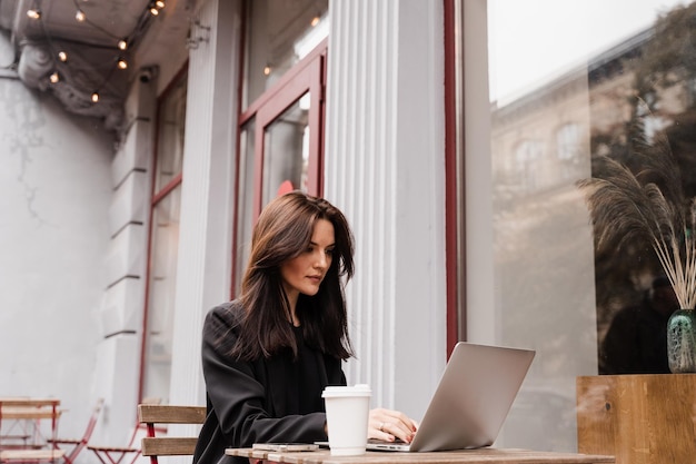 Atrakcyjna dziewczyna programista z laptopem pracuje online poza kawiarnią na tarasie Pewna siebie młoda bizneswoman pracuje online z zespołem na laptopie nad projektem startowym