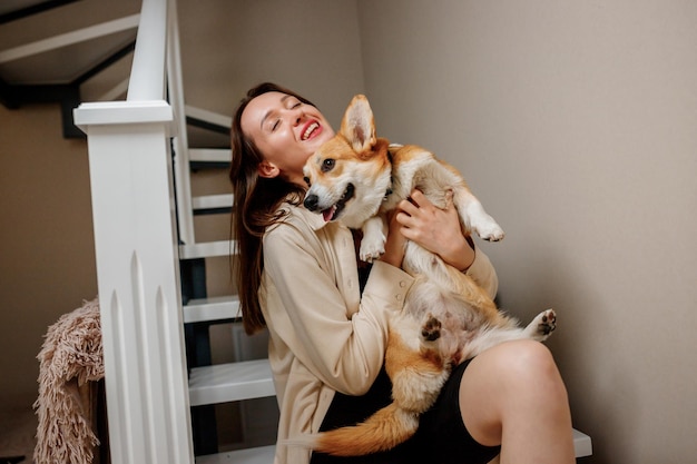Atrakcyjna dziewczyna przytula się i bawi się z psem corgi w domu Welsh Corgi Pembroke z jego właścicielką kobieta siedzi na schodach