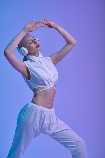 Atrakcyjna futurystyczna kobieta w białych strojach i nowoczesnych słuchawkach, niebieskie tło. Seksowna kobieta w stylu wirtualnej rzeczywistości, technologia przyszłości, futuryzm