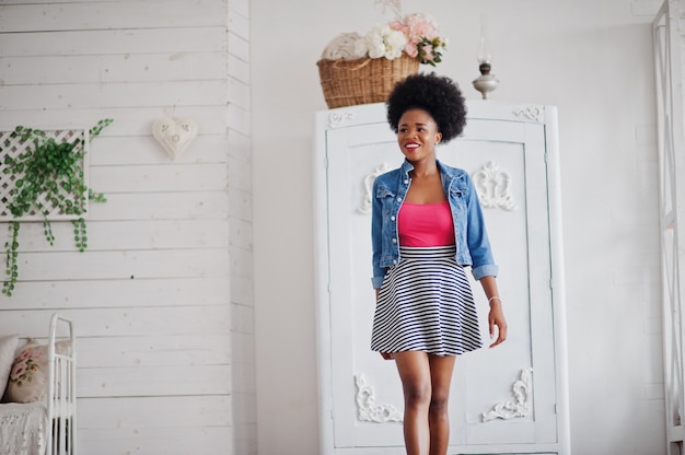 Atrakcyjna kobieta Afroamerykanów z afro włosów nosić na spódnicy i dżinsowej kurtce, pozuje w białym pokoju. Modny czarny model.