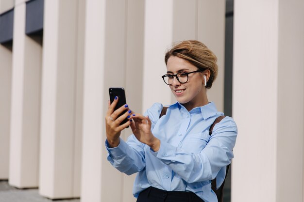 Atrakcyjna kobieta hipster w swobodnym stroju i okularach za pomocą smartfona do czatu wideo. Młoda blondynka stoi w pobliżu biurowca i rozmawia online.