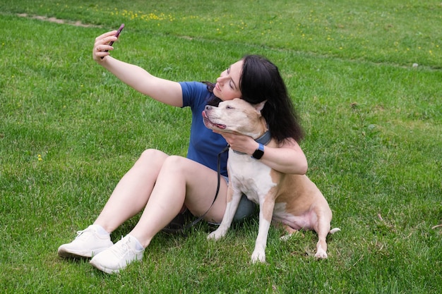 Atrakcyjna kobieta o ciemnych długich włosach siedzi na trawniku z psem i robi sobie selfie przez telefon