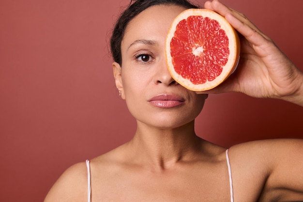 Atrakcyjna kobieta o idealnie zdrowej skórze trzyma przed oczami grejpfruta, zakrywając nim połowę swojej pięknej twarzy. Stosowanie naturalnych organicznych składników roślinnych w kosmetykach do pielęgnacji skóry