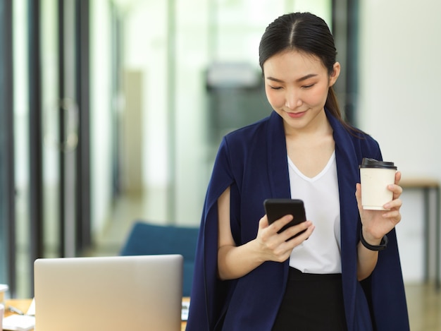 Atrakcyjna kobieta pije kawę i używa inteligentnego telefonu komórkowego, stojąc w biurze, wiadomość młodego przedsiębiorcy przedsiębiorcy z dostawcą biznesowym w biurze