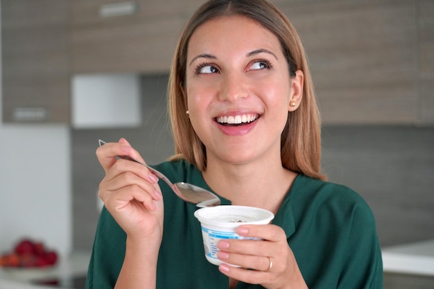 Atrakcyjna kobieta trzyma w dłoni jogurt i uśmiecha się patrząc z boku w pomieszczeniu