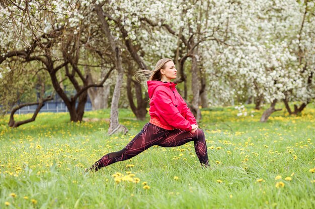 Atrakcyjna kobieta ubrana w różowy bombowiec robi ćwiczenia fitness, rozciągając się na świeżym powietrzu w parku przed kwitnącymi drzewami