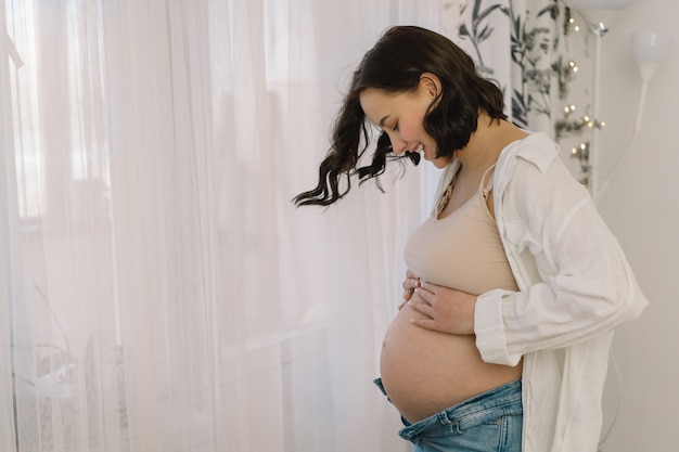 Atrakcyjna kobieta w ciąży stoi przy oknie i trzyma jej brzuch.