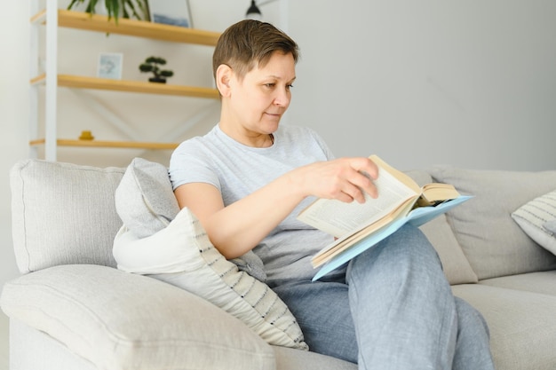 Atrakcyjna kobieta w średnim wieku z uroczym uśmiechem siedząca na kanapie w salonie, ściskając książkę