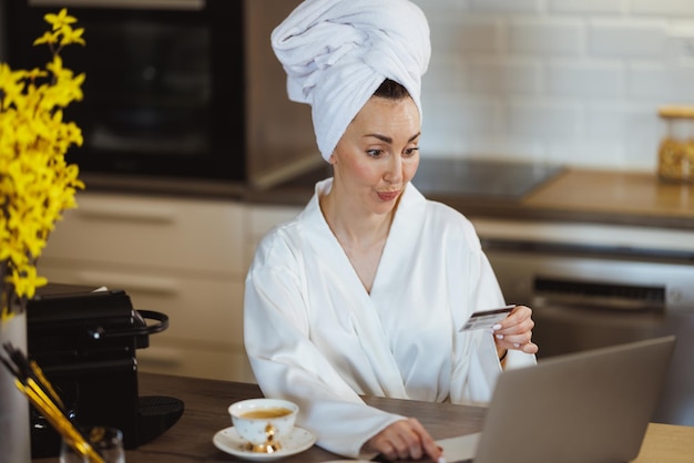 Atrakcyjna kobieta w szlafroku robi zakupy online na laptopie, pijąc poranną kawę i relaksując się w domu.